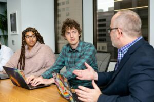 Minimalist editorial illustration in stoic style depicting four professionals seated around a modern conference table, one sharing insights from an open journal while others listen attentively with notebooks. The scene conveys calm collaboration with muted neutral colors, soft natural light, and generous negative space.